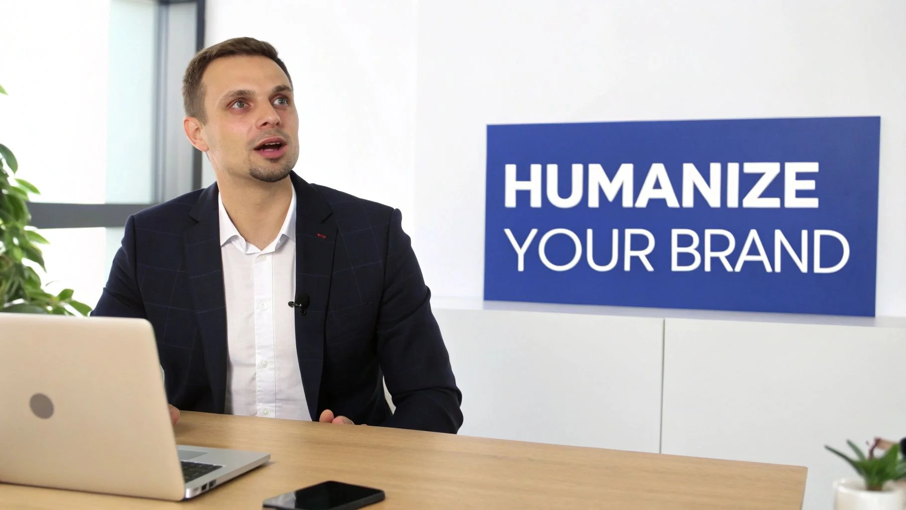Een professionele man in pak spreekt aan een bureau met een laptop, naast een 'Humanize Your Brand' bord.